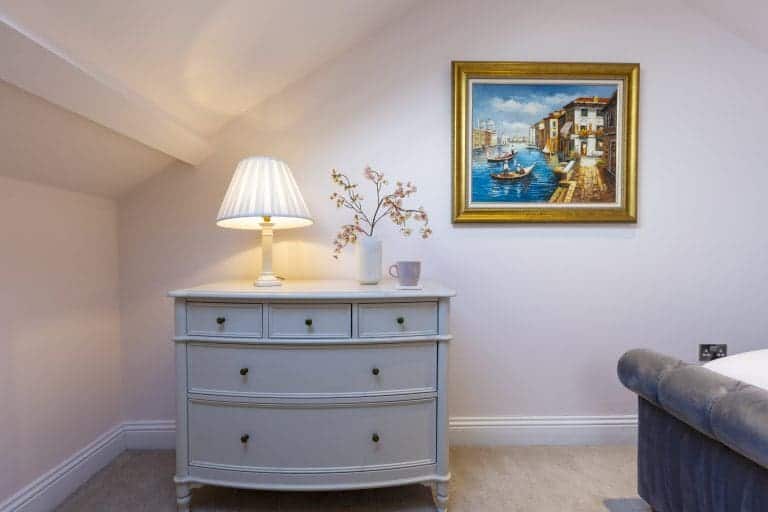 Elegant bedroom corner with a white dresser, lamp, and framed seaside painting.