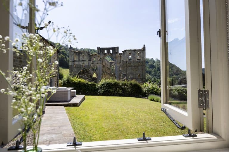 View of Rievaulx Abbey through an open window with a garden in the foreground.