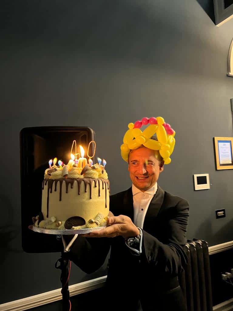 Man in tuxedo holding birthday cake with lit candles and balloons hat.